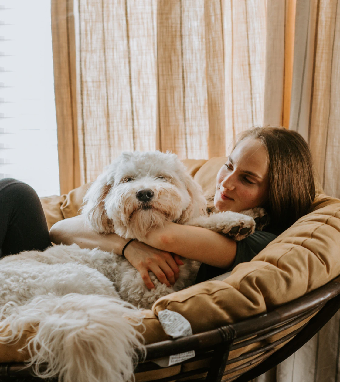 Desktop_About_Us_Our_Purpose A woman sits in a cozy chair, hugging a large, fluffy white dog. They both look relaxed and content, with soft light coming through beige curtains behind them.