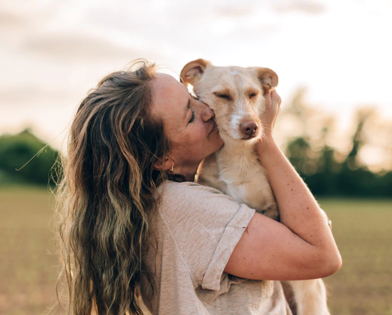 Desktop_Home_Care_Thats A woman with wavy hair hugs and kisses a small, light-colored dog outdoors in a field at sunset.