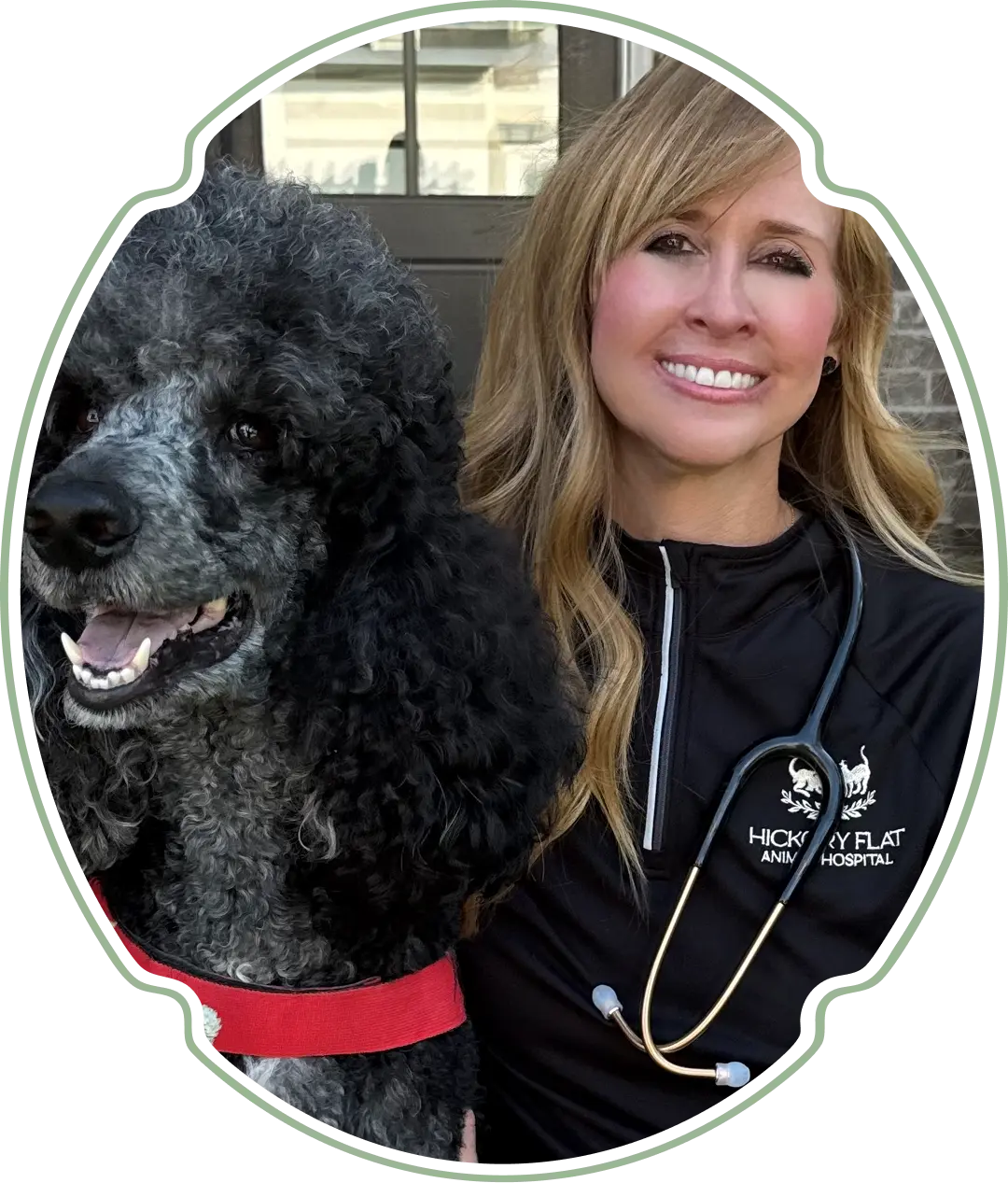 Desktop_Meet_the_Doctors_Maria_Salazar A smiling woman in a black Hickory Flat Animal Hospital jacket poses next to a happy black and gray poodle wearing a red harness. The woman has a stethoscope around her neck.